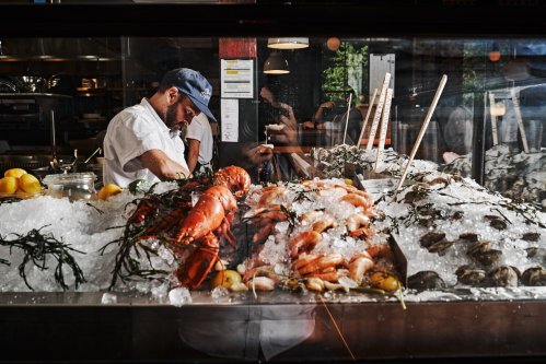 A seafood display with ice, lobsters, shrimp, and oysters, while a chef prepares in the background.