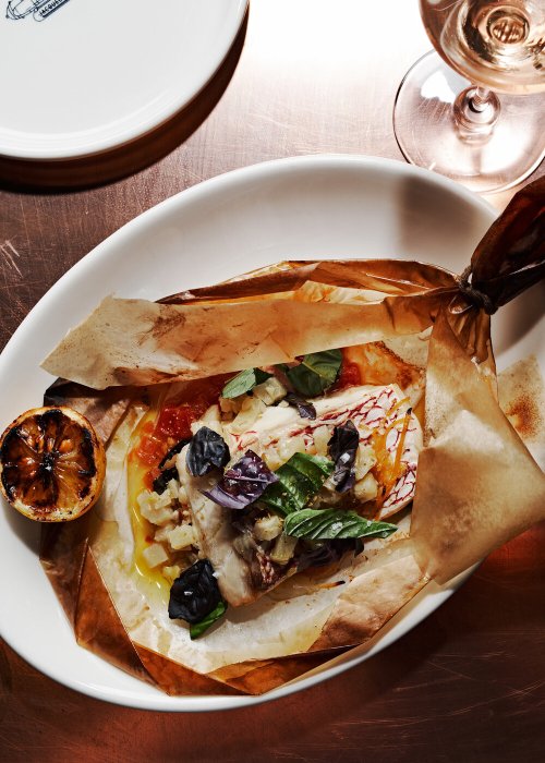 A beautifully plated fish dish in parchment, garnished with herbs, tomatoes, and a charred lemon, with a glass of wine nearby.