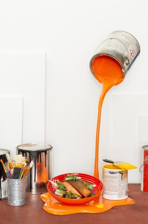 A can of orange paint spills over a bowl of food, creating a vibrant, artistic scene with paint and brushes nearby.