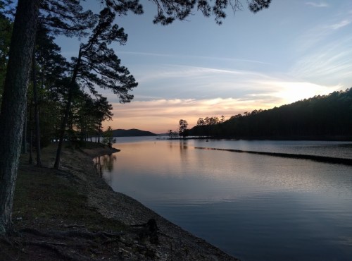 A serene lake at sunset, surrounded by trees and mountains, with calm water reflecting the colorful sky.