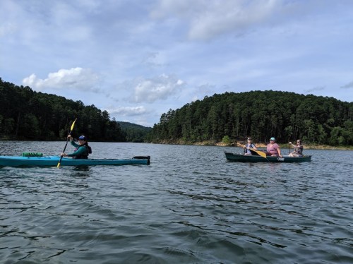 Two kayakers paddle on a calm lake surrounded by lush green trees under a partly cloudy sky.