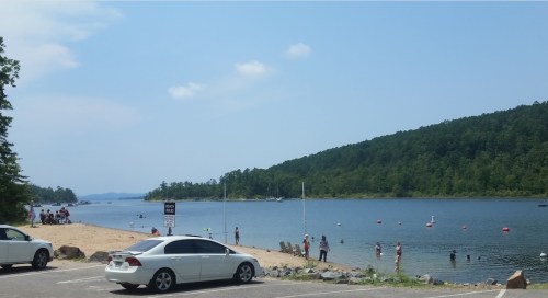 A sunny lakeside scene with people swimming and playing on the beach, surrounded by trees and mountains in the background.