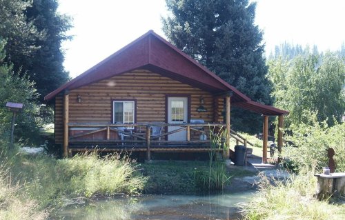 A cozy log cabin with a red roof, surrounded by greenery and a small pond in front.