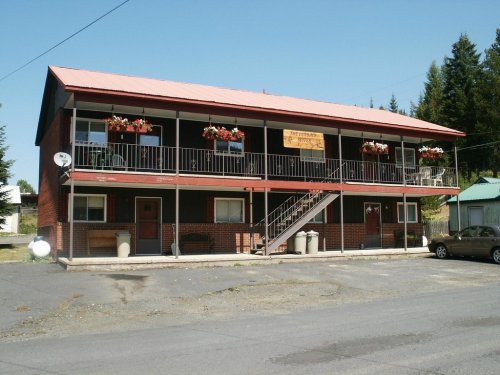 Two-story brick building with a red metal roof, flower boxes, and a staircase, set in a rural area.