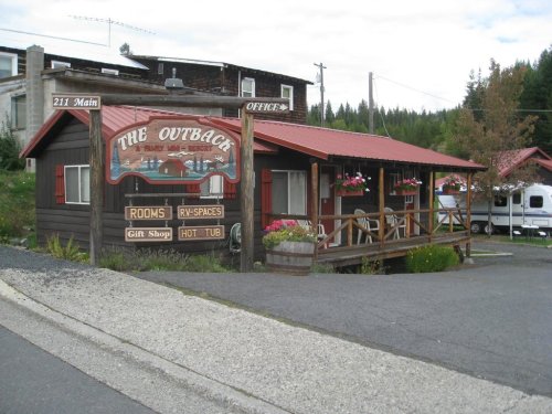 A rustic lodge with a sign reading "The Outback," featuring flower boxes and a hot tub, surrounded by trees.