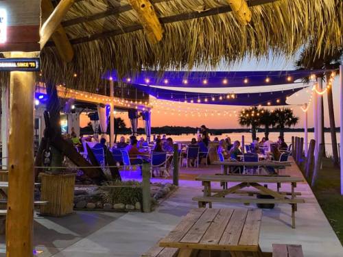 Outdoor dining area with string lights, palm trees, and a sunset view over the water, filled with people enjoying the evening.