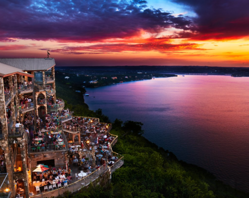 A scenic view of a cliffside restaurant at sunset, overlooking a lake with vibrant colors in the sky.