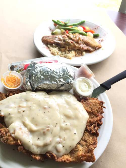 A plate of fried chicken smothered in gravy, with a side of a baked potato and dipping sauce, alongside a vegetable dish.