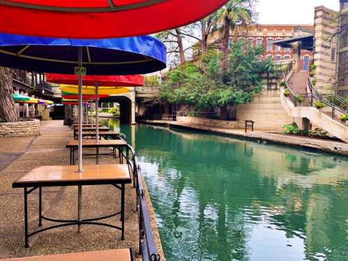 Colorful umbrellas shade empty tables along a serene riverbank, with lush greenery and historic buildings in the background.