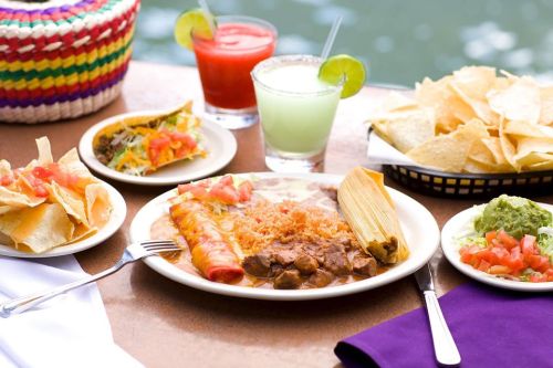A colorful spread of Mexican food including tacos, enchiladas, rice, guacamole, chips, and drinks on a table.