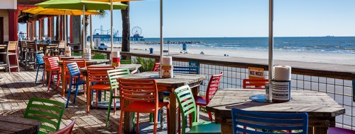 Colorful outdoor seating area with wooden tables overlooking a beach and ocean, with a boardwalk and amusement park in the distance.