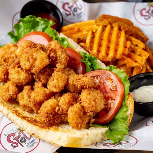A crispy shrimp sandwich with lettuce and tomato, served with sweet potato fries and dipping sauces on the side.