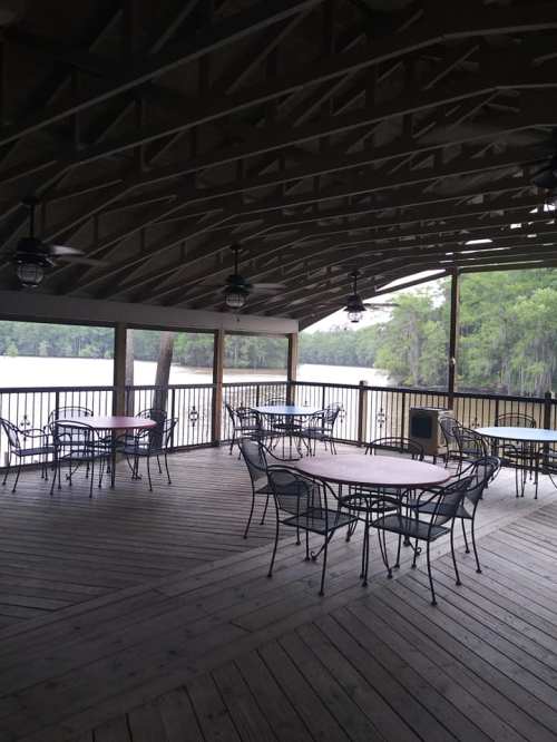 A covered wooden deck with empty tables and chairs overlooking a calm river surrounded by greenery.
