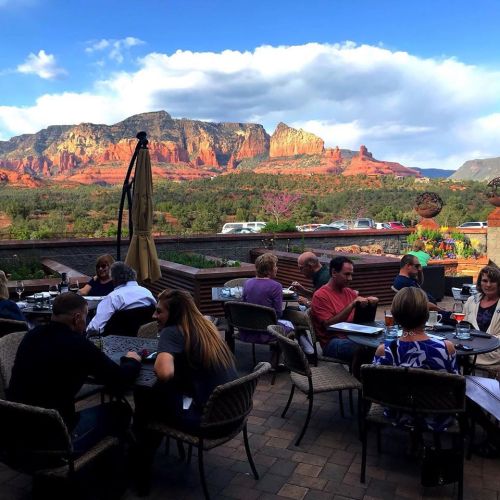 Outdoor dining scene with people at tables, featuring stunning red rock mountains and a blue sky in the background.