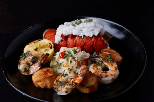 A gourmet seafood dish featuring lobster, shrimp, scallops, and a side of lemon and salad on a black plate.
