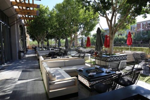 Outdoor dining area with modern furniture, shaded by trees, featuring red umbrellas and a spacious, inviting atmosphere.