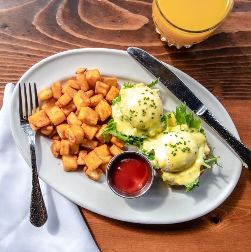 A plate of eggs Benedict with hollandaise sauce, served with crispy potatoes and a side of ketchup.