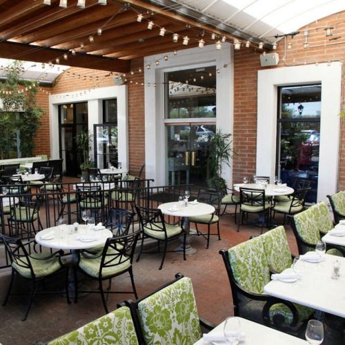 Outdoor dining area with green-patterned chairs, round tables, and string lights under a covered patio.