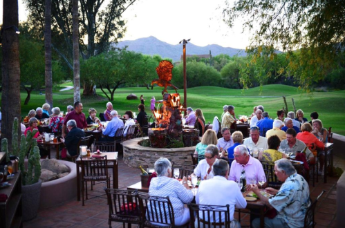 A lively outdoor dining scene with guests enjoying a meal near a fire pit, surrounded by greenery and mountains.