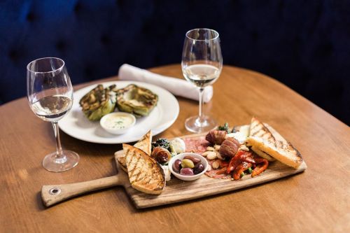 A wooden platter with assorted meats, olives, and grilled bread, accompanied by two glasses of white wine and a plate of artichokes.