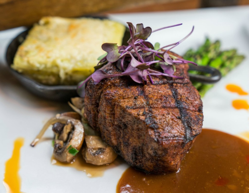 A grilled steak topped with microgreens, served with mushrooms, asparagus, and a side of cornbread.