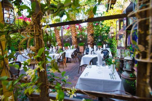 A charming outdoor restaurant patio with white tablecloths, surrounded by greenery and palm trees.