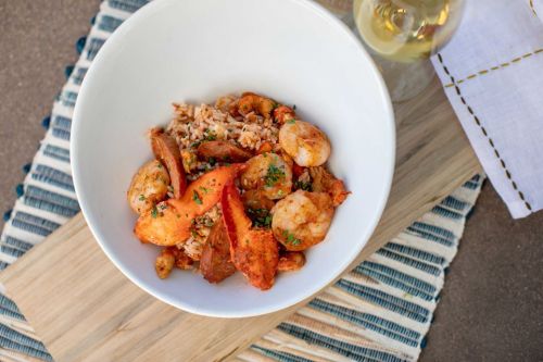 A bowl of shrimp and lobster over rice, garnished with herbs, beside a glass of white wine on a textured surface.
