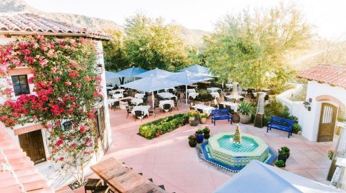 A sunny outdoor dining area with tables, umbrellas, and a fountain, surrounded by lush greenery and colorful flowers.