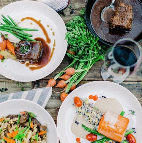 A table spread with three gourmet dishes: glazed meat, salmon with sauce, and pasta with vegetables, garnished with herbs.