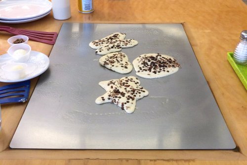 Three star-shaped pancakes with chocolate chips cooking on a griddle, surrounded by plates of butter and syrup.