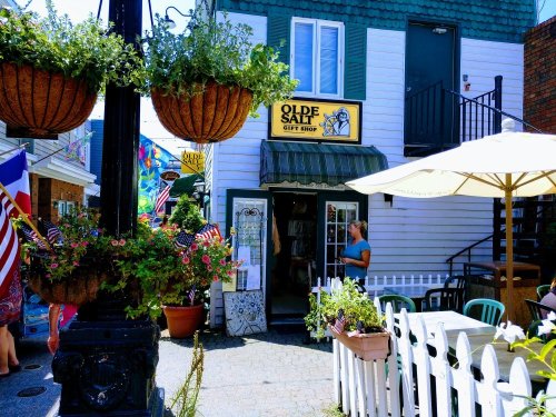 A quaint gift shop with a green awning, surrounded by hanging plants and outdoor seating, in a vibrant market area.