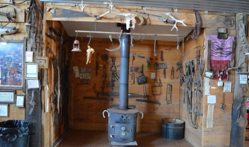 A rustic wooden room featuring a black stove surrounded by various tools and artifacts hanging on the walls.