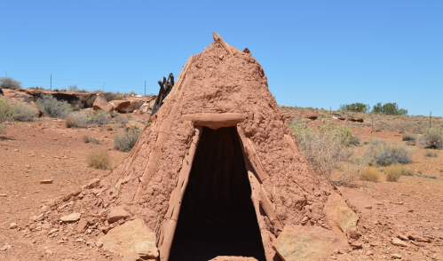 A small, traditional adobe structure with a triangular roof, set in a desert landscape under a clear blue sky.