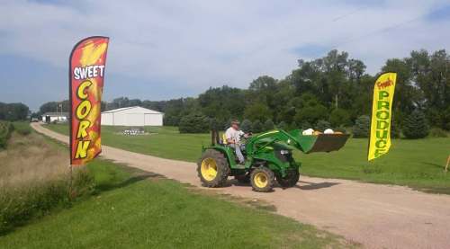 A farmer drives a green tractor on a dirt road, passing colorful signs for sweet corn and produce.