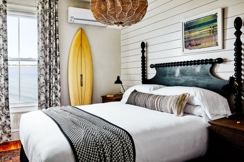 Cozy bedroom with a surfboard, patterned curtains, and a view of the water, featuring a stylish bed and decor.