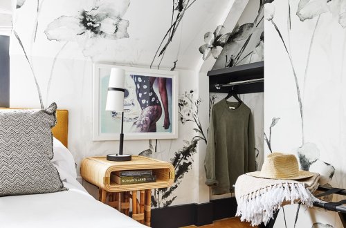 A cozy bedroom with floral wallpaper, a bed with patterned pillows, a lamp, and a closet with a sweater and hat.