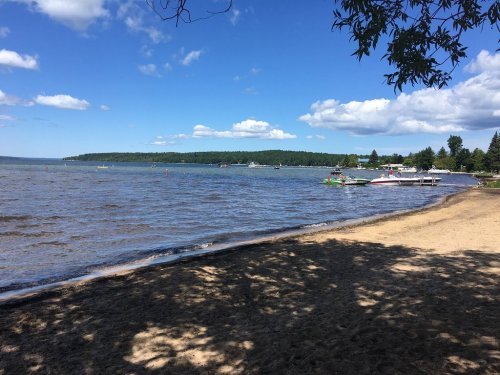 A serene lakeside view with boats on the water, sandy beach, and trees under a blue sky with fluffy clouds.