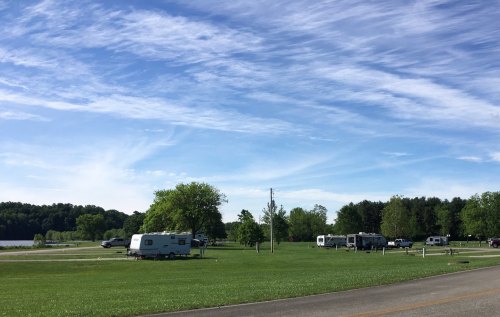 A grassy area with several RVs parked near a lake under a blue sky with wispy clouds. Trees line the background.
