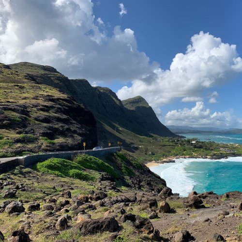 Scenic coastal view with rocky cliffs, a winding road, and turquoise waters under a partly cloudy sky.