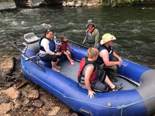 A family sits in a blue raft by a river, preparing for a fishing trip, with trees and a bridge in the background.