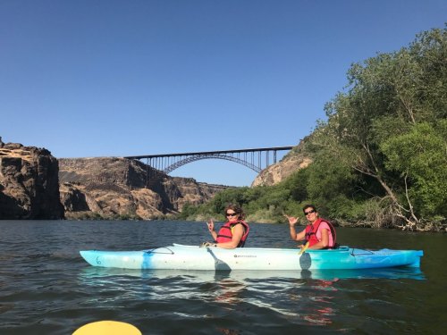 Two people in a kayak on a river, with a bridge and rocky cliffs in the background under a clear blue sky.