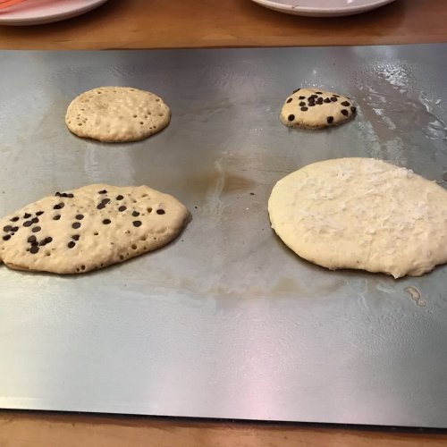 Four pancakes cooking on a griddle, two with chocolate chips and one with shredded coconut.