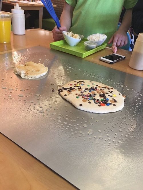 A griddle with two pancakes, one plain and one topped with colorful chocolate candies, and a bowl of ice cream nearby.