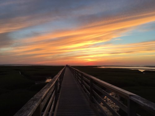 A wooden pier stretches into a vibrant sunset over marshland, with colorful clouds reflecting in the sky.