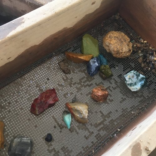 A wooden box containing various colorful gemstones and minerals on a mesh screen.