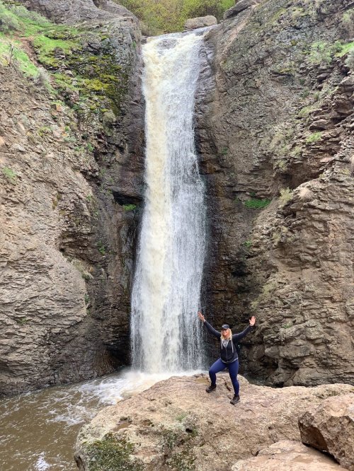 A person stands on a rock with arms raised in front of a tall waterfall surrounded by rocky terrain and greenery.
