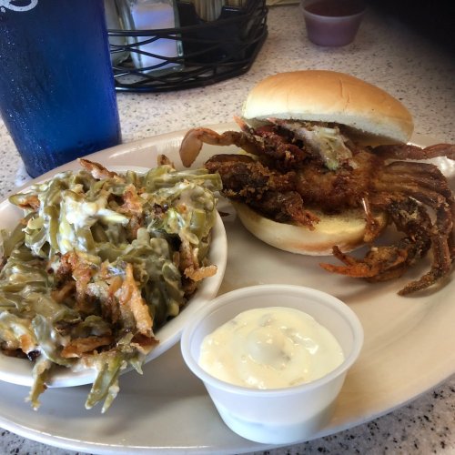 A plate featuring a soft shell crab sandwich, a side of green bean casserole, and a small cup of dipping sauce.