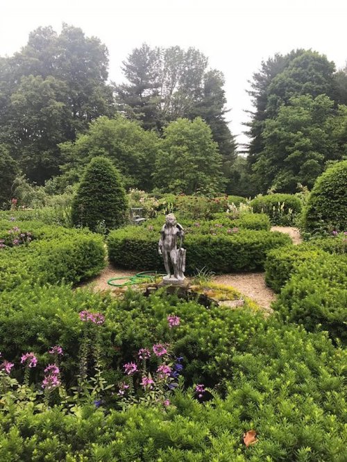 A serene garden with lush greenery, flowering plants, and a central statue surrounded by neatly trimmed hedges.