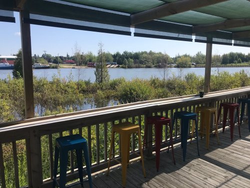 A wooden deck with colorful stools overlooks a calm river surrounded by greenery on a sunny day.