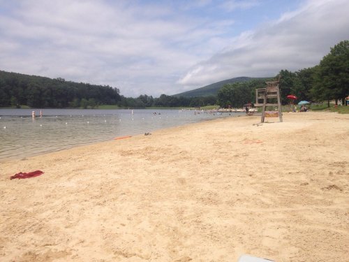 A sandy beach by a calm lake, with trees and mountains in the background, and people enjoying the water.